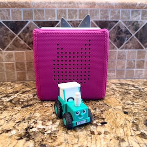 May include: A fuchsia-colored audio player with a perforated speaker and silver ears, next to a teal toy tractor with a white roof. The tractor has a smiling face and the text "TED 1" on the front.