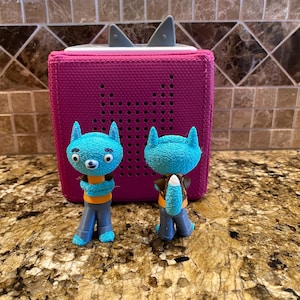 May include: A magenta-colored audio player with two blue cat-like figurines. The audio player has a speaker grill and two gray fin-shaped accents. The figurines are blue with orange and gray accents, resembling cartoon characters.