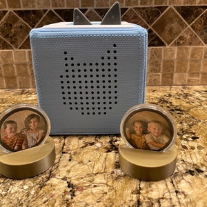 May include: A light blue audio player with shark fin-shaped accents and a speaker. Two photo displays with circular frames and gold-colored bases are in front of the player. Each display holds a photo of children.