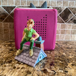 May include: A fuchsia audio player with a Peter Pan figurine on a miniature rooftop. The figurine is wearing a green outfit. The audio player has a speaker and two grey ears.