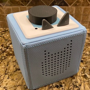 May include: A light blue cube-shaped speaker with a black knob and two gray buttons on top. The speaker has a grid of small holes on the front side.