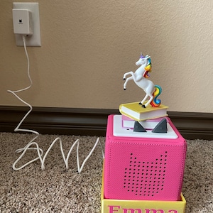 以下が含まれることがあります： 黒いスピーカーグリルと、その上に置かれた白いユニコーンの置物があるピンクのスピーカー。スピーカーは黄色いベースに「Emma」という名前が印刷されています。