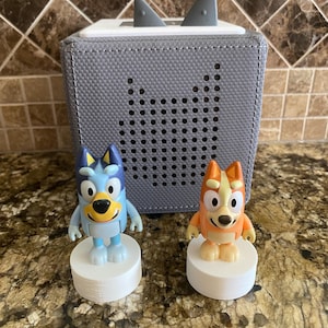 May include: Two small blue and orange cartoon dog figurines stand on white bases in front of a gray speaker with a white top and black ear-like protrusions.