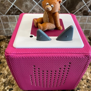 May include: A pink fabric cube speaker with a brown teddy bear sitting on top. The speaker has two gray ear-like buttons on the top and a row of small holes on the front.