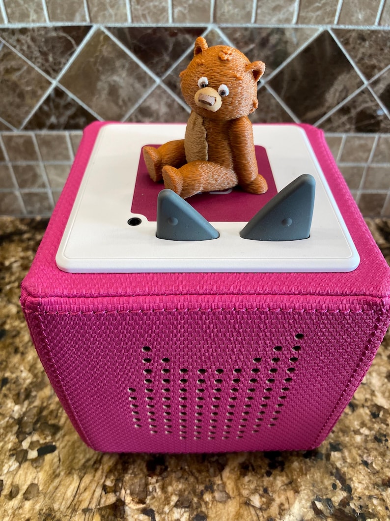May include: A pink fabric cube speaker with a brown teddy bear sitting on top. The speaker has two grey ear-like buttons on the top and a row of small holes on the front.