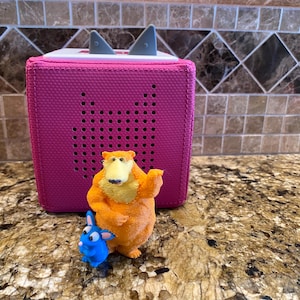 May include: A magenta audio player with a speaker grill and two grey ears on top. In front of the player are two small figurines: an orange bear and a blue rabbit. The objects are on a speckled countertop.