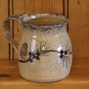 May include: A beige ceramic mug with a brown pine branch and blue berries design. The mug has a speckled glaze and a rounded handle.