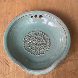 May include: A turquoise ceramic spoon rest with a floral design in the center. The spoon rest has a small indentation for the spoon handle and three small holes near the rim. The rim has a brown accent.