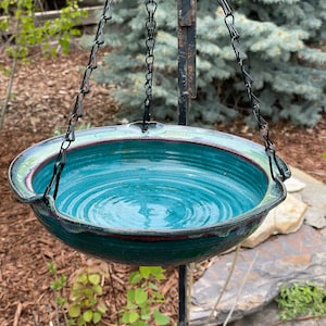 May include: A teal blue ceramic bird bath with a dark red rim hangs from a black chain. The bird bath is suspended from a black metal pole.