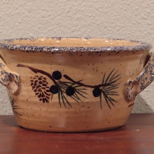 May include: A beige ceramic bowl with two handles, featuring a pine branch and pine cone design in brown and blue.