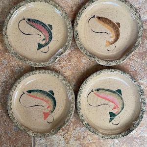 May include: Set of four ceramic plates with a speckled brown glaze. Each plate features a hand-painted design of a trout with a fishing line.