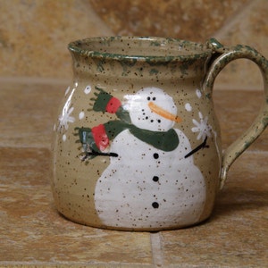 May include: A ceramic mug with a hand-painted snowman design. The snowman has a carrot nose, black coal eyes and mouth, and wears a green and red scarf. White snowflakes are scattered around the snowman on the beige mug.