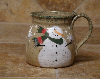 May include: A ceramic mug with a hand-painted snowman design. The snowman has a carrot nose, black coal eyes and mouth, and wears a green and red scarf. White snowflakes are scattered around the snowman on the beige mug.