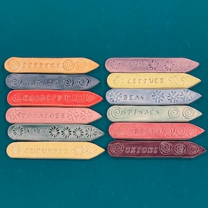 May include: A set of 10 colorful ceramic plant markers with different vegetable names, including peppers, radish, carrots, potatoes, kale, cucumbers, peas, lettuce, beans, and spinach. The markers are decorated with swirls and flowers.