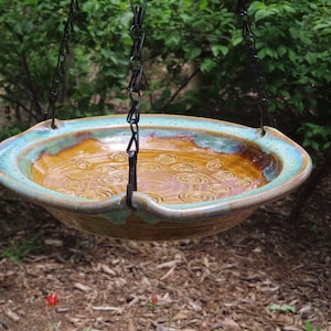 May include: A brown and teal ceramic bird bath with a swirling pattern hangs from a black chain. The bath is suspended over a bed of brown wood chips.