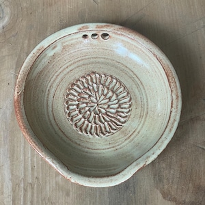 May include: A handmade, light green ceramic grater bowl with a textured floral design in the center. The bowl has a wide rim with a reddish-brown edge and three small holes. The bowl is designed for grating ingredients.