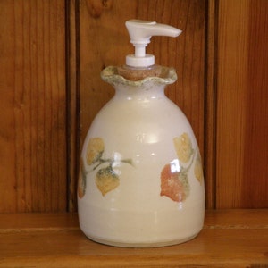 May include: A white ceramic soap dispenser with a leaf design in shades of green, yellow, and orange. The dispenser has a white pump and a cork stopper.