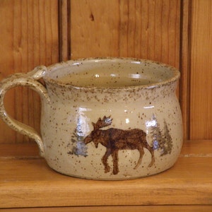 May include: A beige ceramic mug with a brown moose design. The mug has a speckled glaze and a rounded handle.