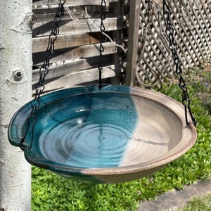 May include: A ceramic bird bath with a teal and beige glaze hangs from a black chain. The bath is round and has a slight lip around the edge.