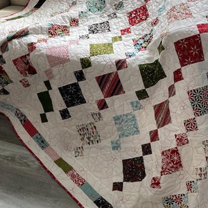 Peut inclure: Une courtepointe blanche avec un motif patchwork rouge, vert et bleu. La courtepointe est pliée et drapée sur un fauteuil beige. La courtepointe a un fond blanc avec un motif floral et une bordure rouge.