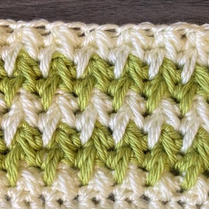 May include: Close-up of a crocheted fabric with a textured pattern. The design features alternating rows of white and light green yarn, creating a zig-zag effect. The stitches are tightly woven, showcasing the handmade quality.