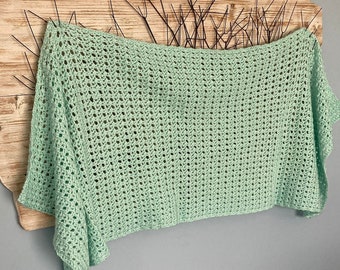 May include: A light green crocheted blanket with a delicate open-stitch pattern. The blanket is draped over a wooden surface with a decorative metal design.