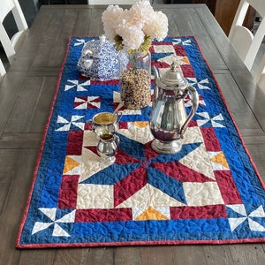 May include: A blue, red, and white quilted table runner with a star pattern. A silver teapot, sugar bowl, and creamer are on the runner, along with a vase of white flowers and a bowl of grain.