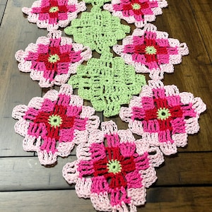 Puede incluir: Un camino de mesa tejido a crochet con un diseño floral rosa, rojo y verde. El camino de mesa está hecho de hilo y tiene un patrón delicado y de encaje.