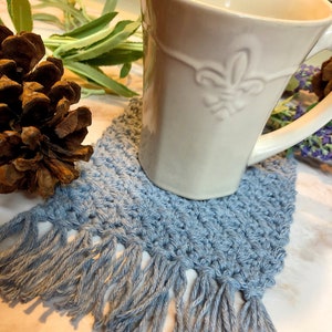 May include: A blue crocheted coaster with fringe, featuring a white ceramic mug with a decorative design. The coaster is sitting on a white marble surface.