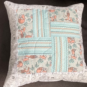 May include: A light blue and white patterned pillow with a patchwork design featuring foxes and trees. The pillow has a white border with a black and white pattern of foxes, trees, and other woodland creatures.