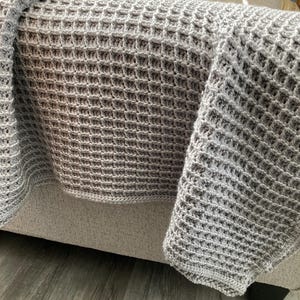 May include: A gray, hand-crocheted blanket with a waffle-like texture draped over a beige sofa. The blanket's intricate pattern and soft color create a cozy and inviting aesthetic. The blanket is made of a soft yarn.