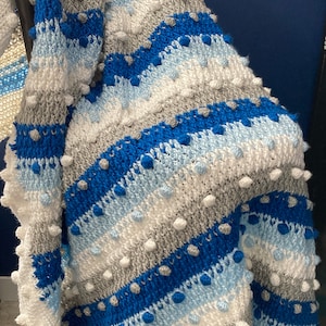 May include: A striped crocheted blanket with blue, white, and gray stripes. The blanket has a textured, bobbly surface.