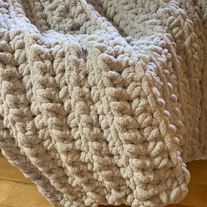 Peut inclure: Une couverture en tricot &eacute;pais et doux de couleur gris avec un motif textur&eacute;. La couverture est pli&eacute;e en trois et repose sur un sol en bois.