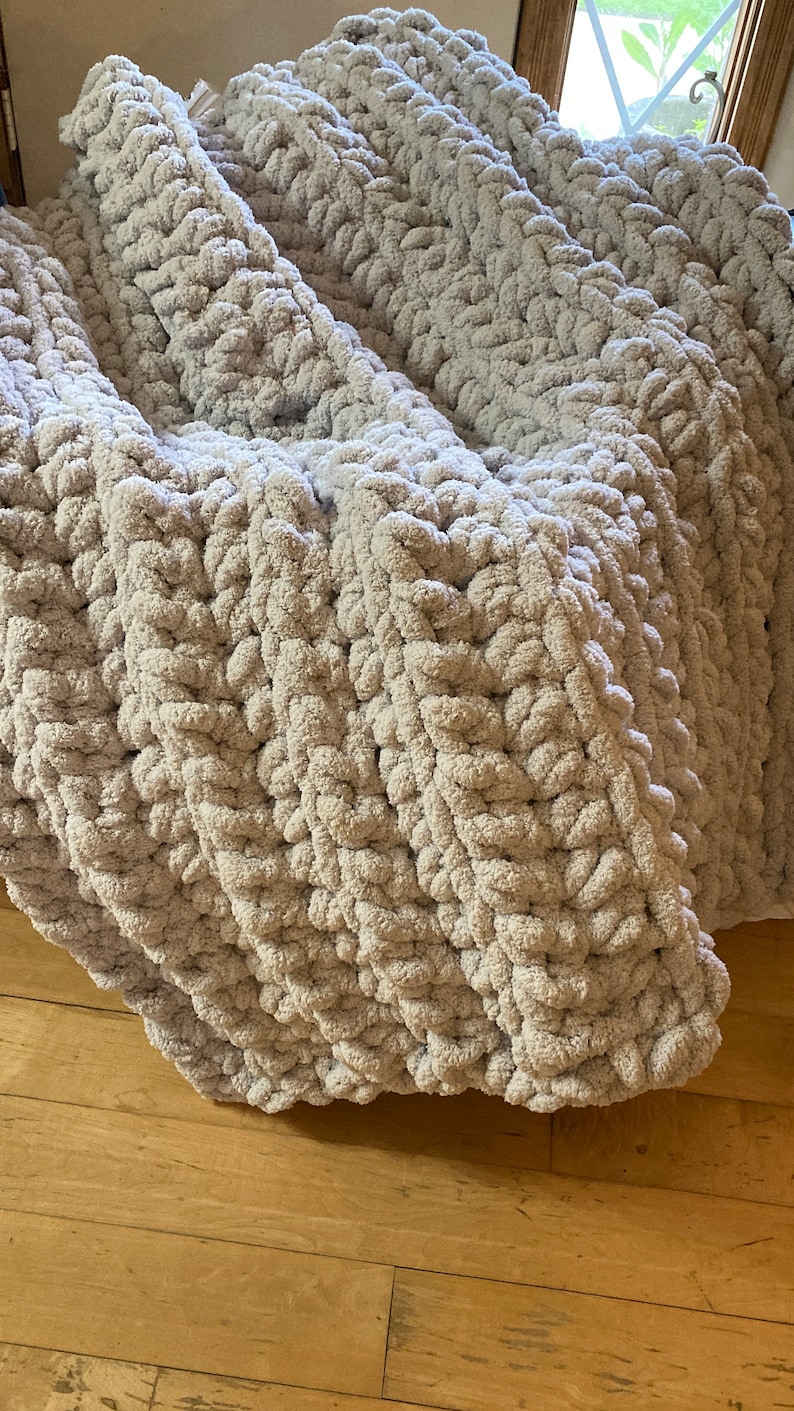 Peut inclure: Une couverture en tricot &eacute;pais et doux de couleur gris avec un motif textur&eacute;. La couverture est pli&eacute;e en trois et repose sur un sol en bois.