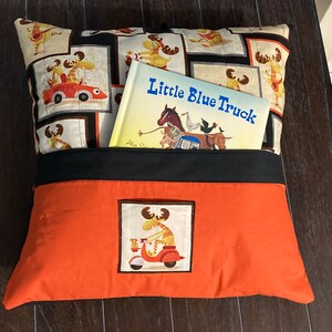 May include: Orange and black pillow with a pocket for a book. The pillow has a moose design on the front and a moose design on the back. The book is titled "Little Blue Truck".
