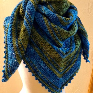 May include: A handmade, triangular crochet shawl in shades of blue and green. The shawl is draped over a mannequin, showcasing its textured stitch pattern and scalloped edges. The color combination creates a visually appealing and cozy accessory.