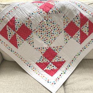 May include: A white quilt with a geometric pattern featuring red squares and triangles. The quilt has a colorful floral pattern in the center and along the edges. The quilt is folded over a beige sofa.