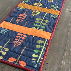 May include: A quilted table runner with a navy blue background and a pattern of colourful leaves. The runner has a red border and is made of fabric.