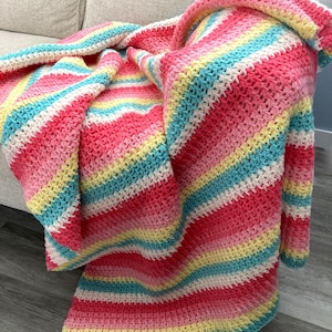 Könnte beinhalten: Eine handgehäkelte Decke mit einem gestreiften Muster in Rosa, Rot, Gelb, Blau und Weiß. Die Decke ist über ein Sofa drapiert, wodurch ihre Textur und ihr Design hervorgehoben werden. Die Decke wirkt weich und gemütlich.
