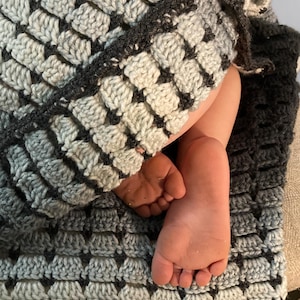 Op de afbeelding: Een handgemaakte gehaakte deken in grijstinten en wit, met een textuur, geometrisch patroon. De deken is over een persoon gedrapeerd, met hun blote voeten zichtbaar. Het ontwerp van de deken creëert een gezellig en uitnodigend gevoel.