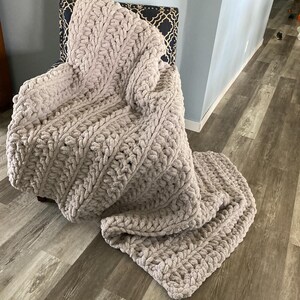 Peut inclure: Une couverture en tricot moelleuse et &eacute;paisse de couleur beige clair. La couverture est drap&eacute;e sur une chaise et pr&eacute;sente un motif textur&eacute; en tricot torsad&eacute;.