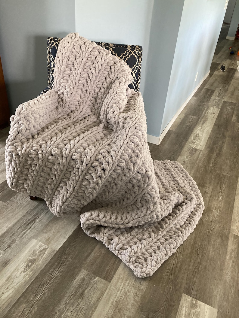 Peut inclure: Une couverture en tricot moelleuse et &eacute;paisse de couleur beige clair. La couverture est drap&eacute;e sur une chaise et pr&eacute;sente un motif textur&eacute; en tricot torsad&eacute;.