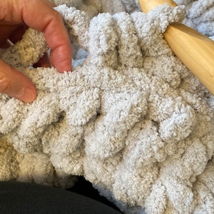 Peut inclure: Gros plan sur une personne crochetant une couverture moelleuse de couleur gris clair avec un crochet en bois. La couverture est faite avec un fil &eacute;pais et a un design textur&eacute; et bouffant.