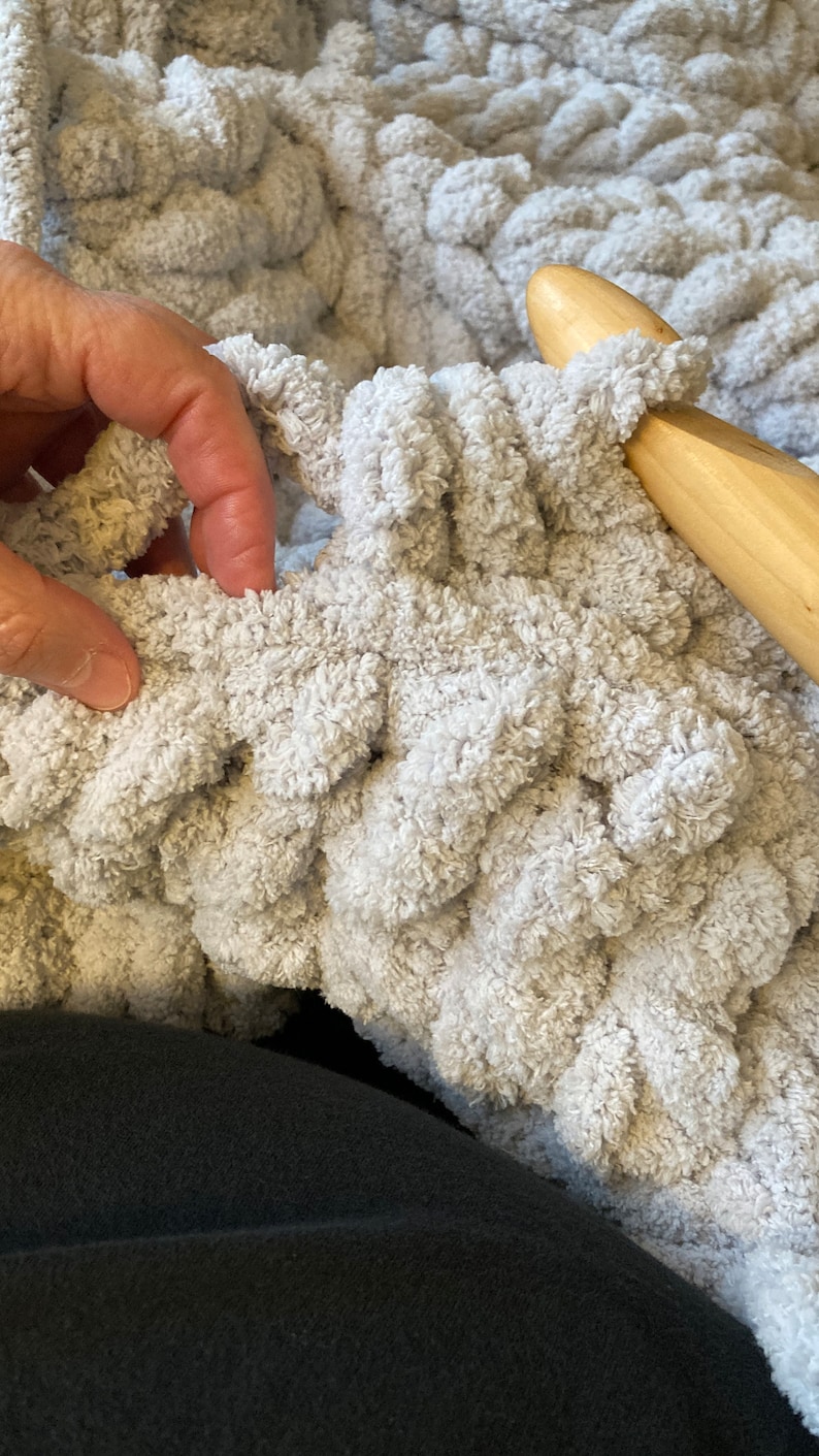 Peut inclure: Gros plan sur une personne crochetant une couverture moelleuse de couleur gris clair avec un crochet en bois. La couverture est faite avec un fil &eacute;pais et a un design textur&eacute; et bouffant.