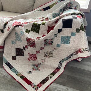 May include: A white quilt with a red, green, and blue patchwork design. The quilt is folded and draped over a white couch.