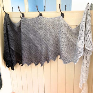 May include: Several crocheted shawls in shades of gray and white hang from hooks on a white wall. The shawls have a scalloped edge and a delicate, open weave design. The shawls are displayed in a row, showcasing their varying colors and textures.