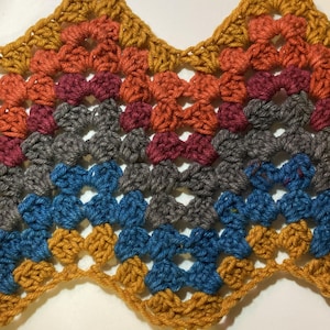 Op de afbeelding: Een close-up van een gehaakt weefsel met een golvende rand. Het weefsel is gemaakt met garen in verschillende kleuren, waaronder oranje, rood, grijs, blauw en geel.