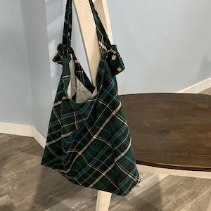 May include: A green, black, and white plaid tote bag hangs on a white chair. The bag has a large capacity and is made of a soft fabric. The straps are knotted at the top and have gold buttons. The bag is a stylish accessory.
