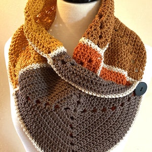 May include: A crocheted cowl with a button closure. The cowl is made in shades of brown, orange, and yellow. The cowl is displayed on a white mannequin.