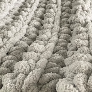 Peut inclure: Une couverture en tricot moelleuse, &eacute;paisse et grise avec un motif tiss&eacute;. La couverture est faite de fil &eacute;pais et a une texture duveteuse.
